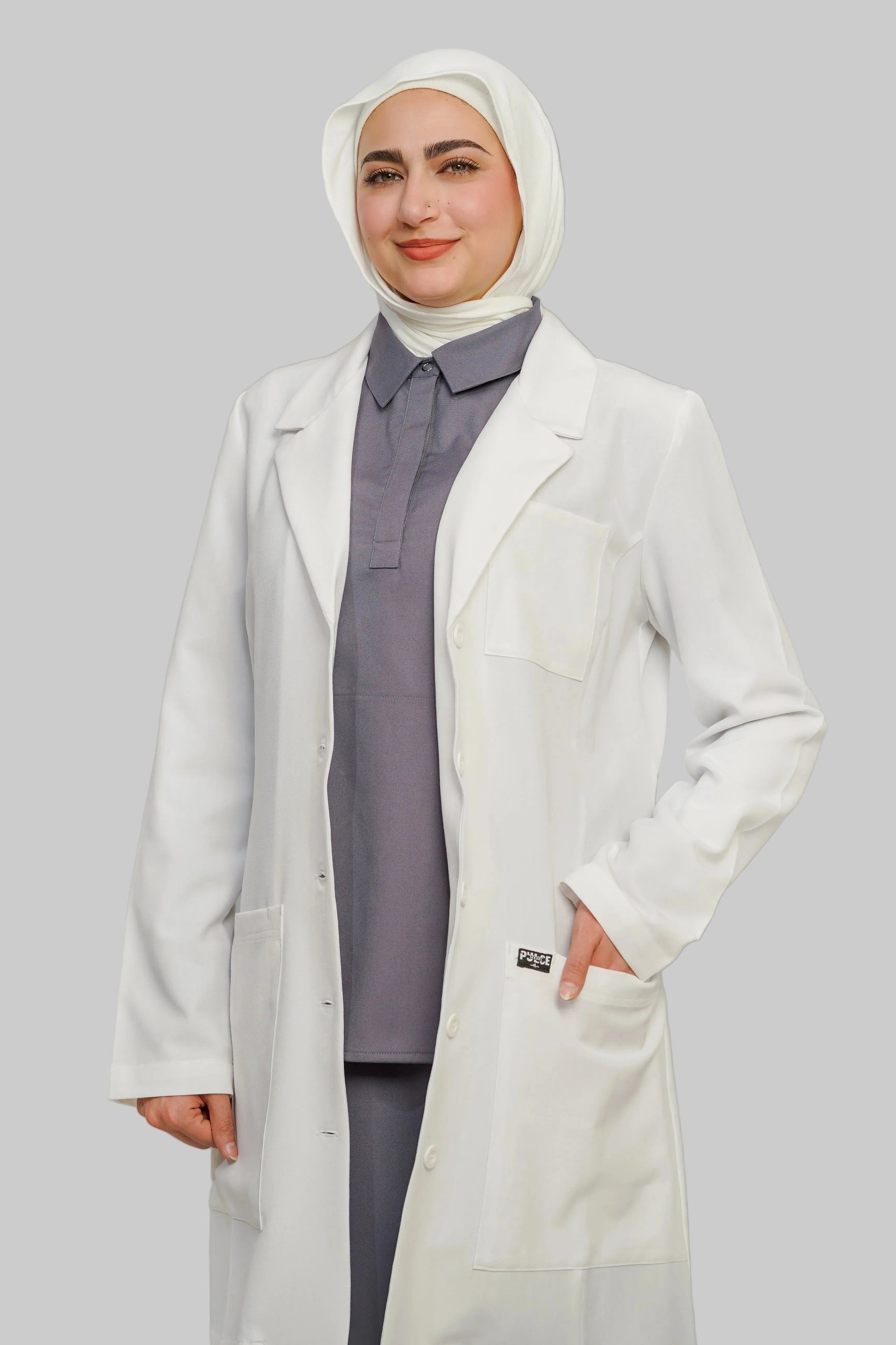 Person wearing a white lab coat over a gray outfit with a headscarf on a plain background