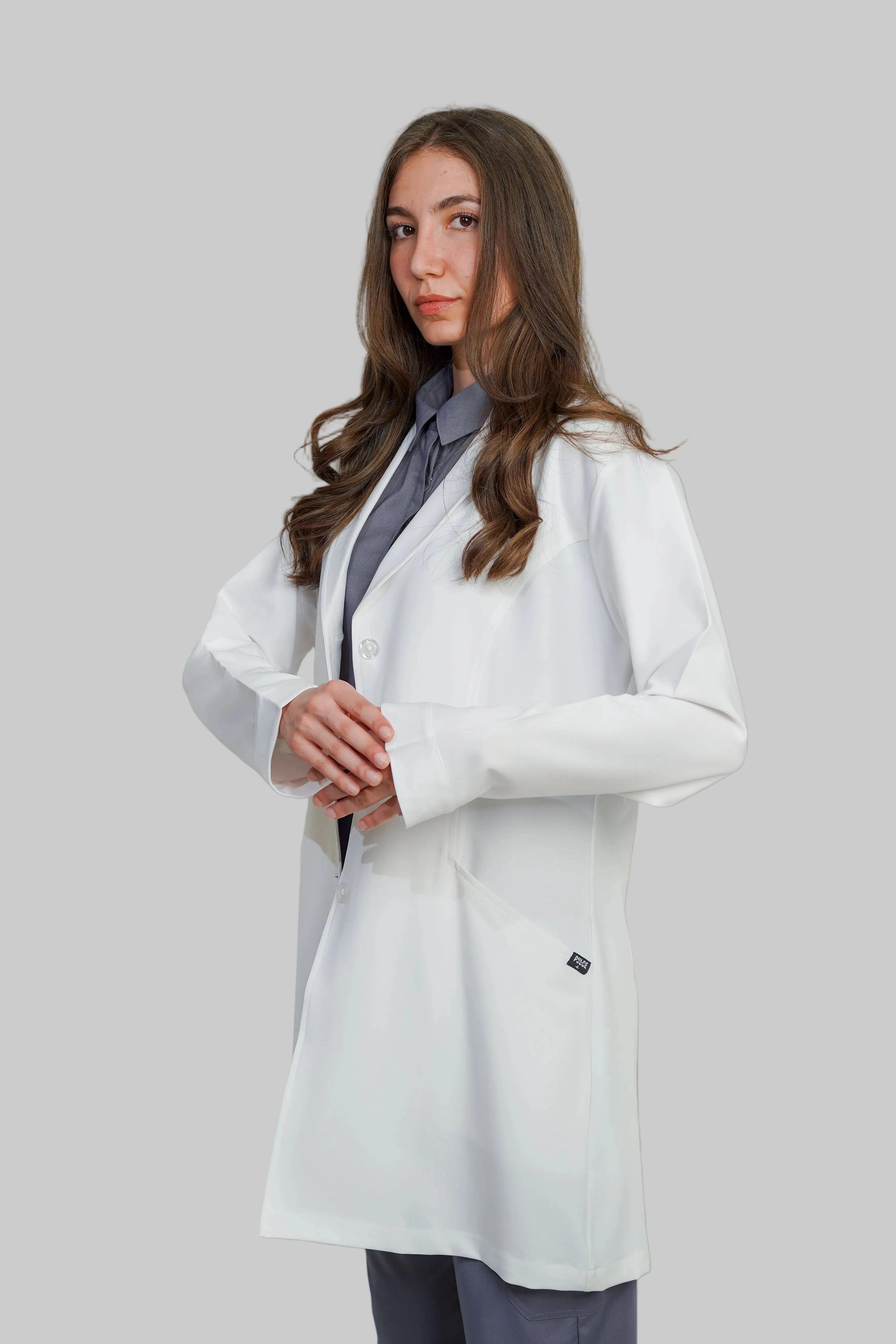 Woman wearing a white lab coat over a gray outfit against a plain background