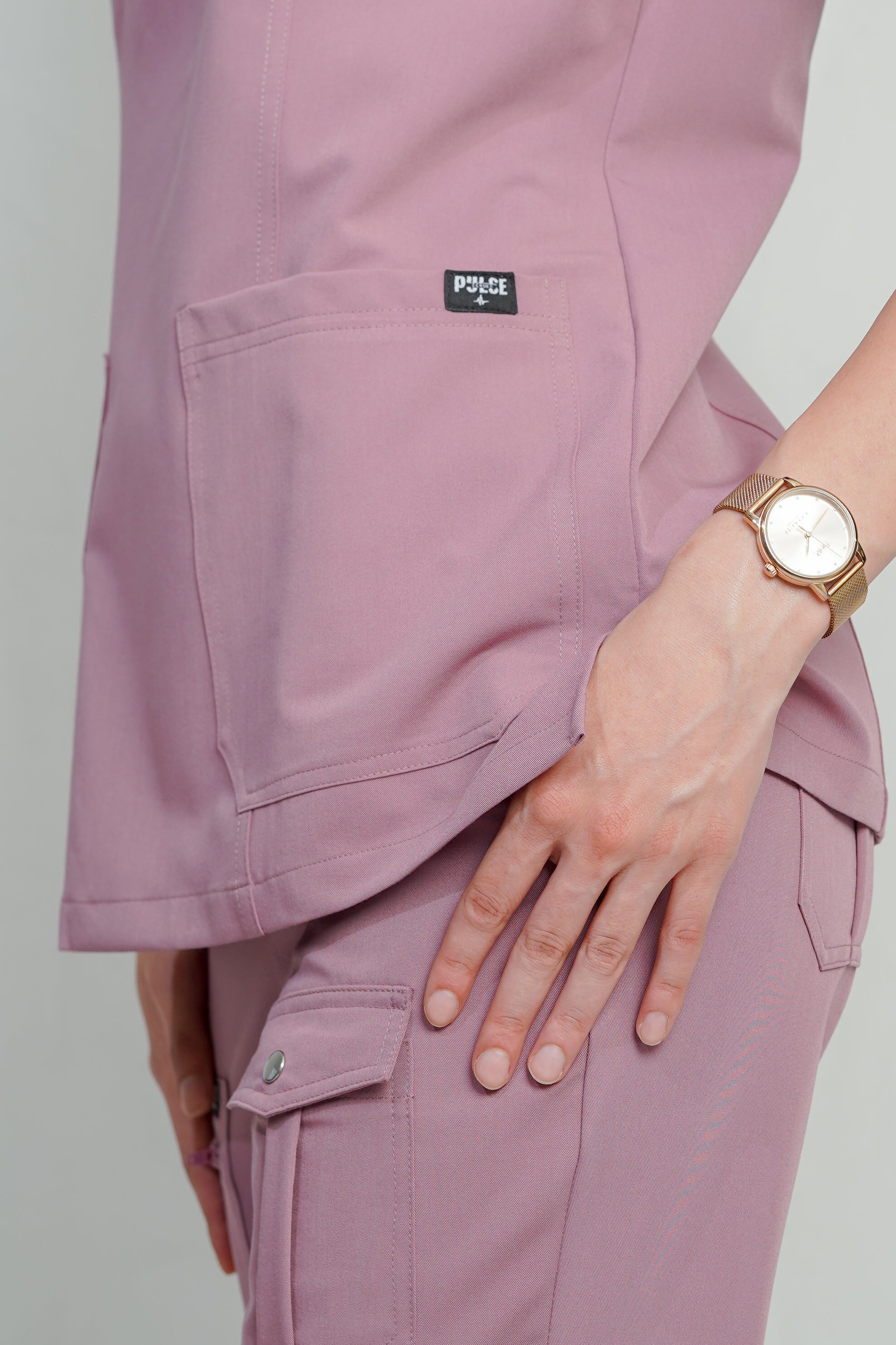 Person wearing a light purple scrub set with a visible brand logo, holding a wristwatch.